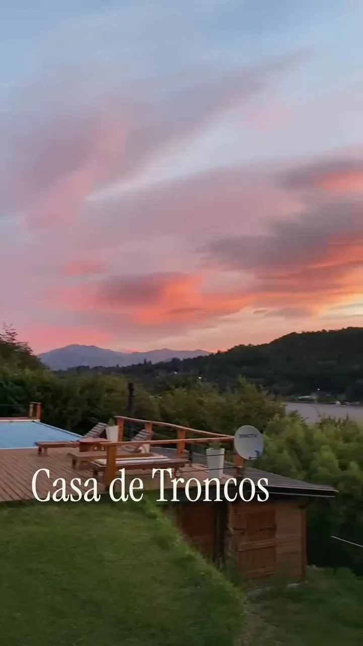 Un recorrido desde la Casa de Troncos y su jardín hacia el lago y el cielo rosado. En pantalla: Casa de Troncos. Entre el bosque y el lago.
