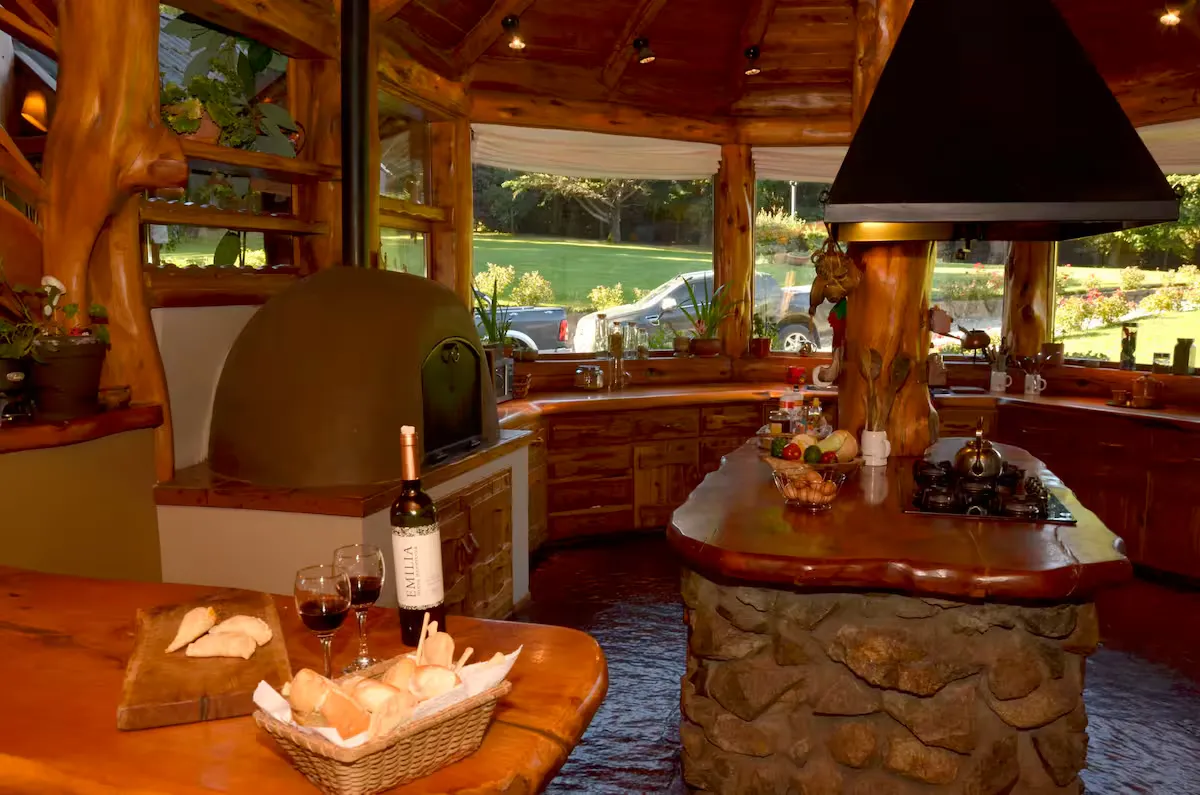 Cocina circular de troncos con horno de barro, isla de piedra y vino sobre la mesa