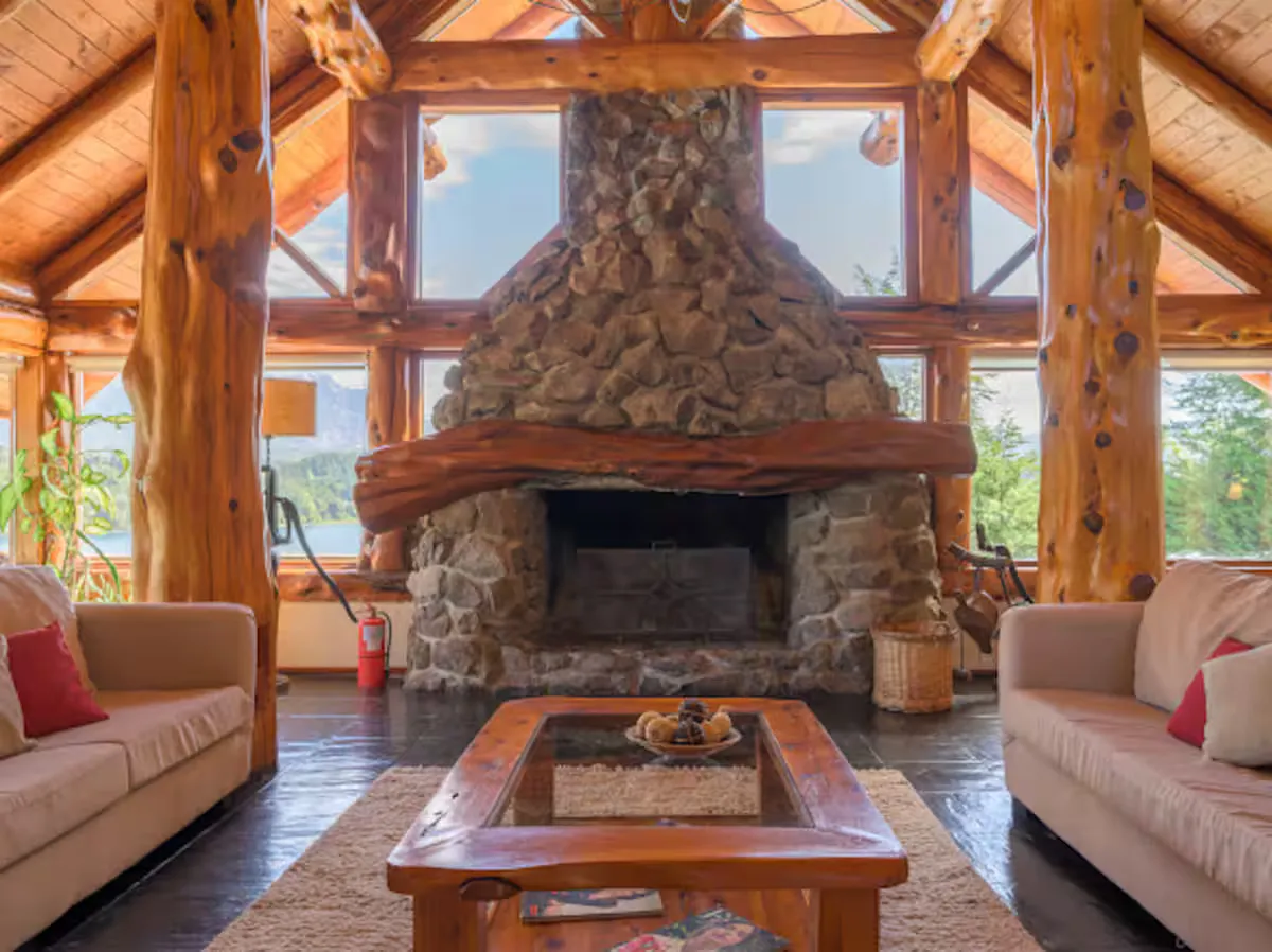 Living de troncos con chimenea de piedra central y ventanales altos